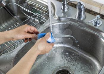 Horizontal photo of female hands washing a small knife with a soapy sponge with kitchen sink and running faucet in background