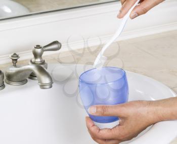 Closeup Photo of female hands putting tooth brush in blue cup with white bathroom sink and faucet in background 