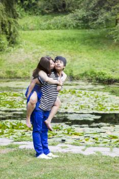 Vertical photo of young adult couple, woman riding on boyfriend back and kissing him, with pond in background 
