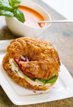 Vertical photo, slightly angled, of chicken salad, tomato, basil, and onion inside croissant bread on white square plate with tomato soup and spoon in bowl all resting on natural stone counter top 