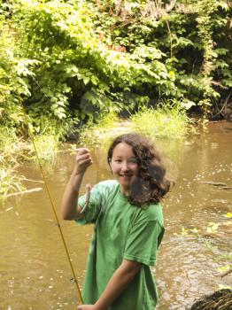 Vertical photo of young girl smiling while holding up small trout with stream and trees in background  

