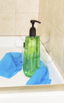 Vertical photo of tall green shower gel bottle, with water drops, wash cloth and a small bottle of hair conditioner with shower stall in background