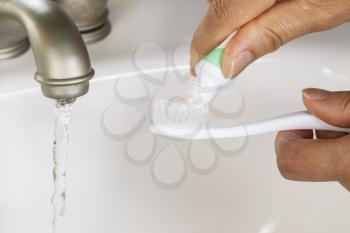 Closeup of horizontal Photo of female hands putting tooth paste on toothbrush with white bathroom sink and running faucet in background 