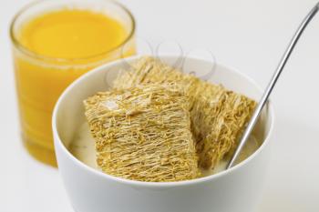 Horizontal photo of natural wheat cereal covered with honey residing inside a white bowl with spoon and milk inside along with a glass of Orange Juice 