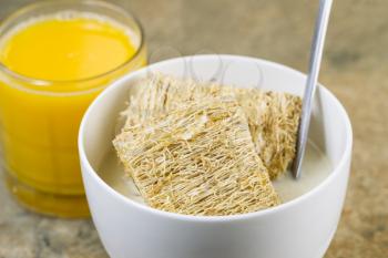 Horizontal photo of natural wheat cereal covered with honey residing inside a white bowl with spoon and milk inside along with a glass of Orange Juice on natural stone counter top

