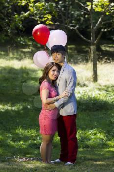 Vertical photo of young adult couple dressed in formal attire holding each other outdoors with several balloons and trees behind them  