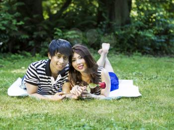 Horizontal photo of young adult couple lying on blanket, looking forward, red rose being held by woman with green grass and trees in background 