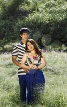 Vertical photo of young adult couple holding each other, with single red rose in hand of woman, while standing in the middle of a bright green field of tall grass and trees in background 