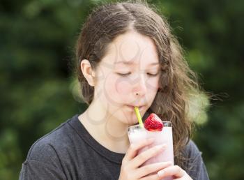 Photo of young girl sipping on a strawberry milkshake during a summer day with blurred out bright green trees in background 