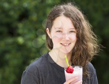 Photo of young girl holding strawberry milkshake outdoors with blurred out bright green trees in background 