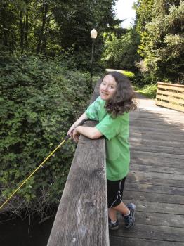 Vertical photo of young girl, looking forward, fishing off of wooden bridge for trout with stream, walk path and trees in background  