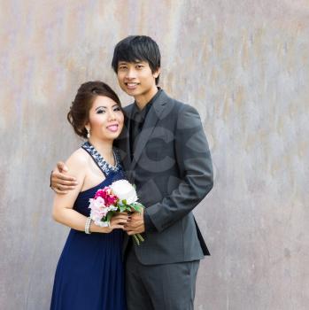 Closeup photo of young adult couple holding a bouquet of flowers, in between them, as they hug each other with rustic metal wall in background 