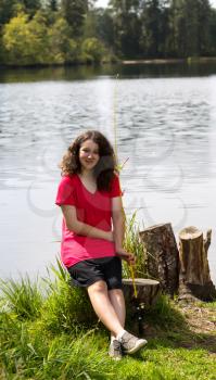 Vertical photo of young girl holding fishing pole and reel, while sitting on a tree stump, with lake and trees in background 