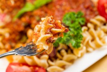 Closeup horizontal photo of freshly cooked pasta in a fork surrounded by cooked curly pasta noodles, tomato sauce, parsley and basil in a white plate 