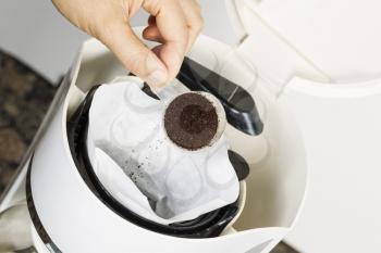 Horizontal angled photo of female hand putting fresh coffee grounds in white bleached coffee filter with coffee maker, and stone kitchen counter top in background 