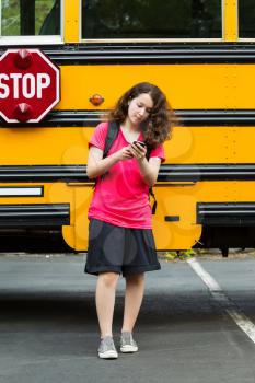 Vertical photo of young girl walking away from school bus, with back pack over her shoulders, while texting on her phone