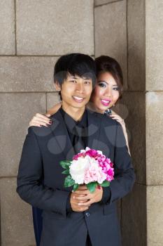 Vertical photo of young adult couple, man holding flowers, looking forward with stone wall in background 