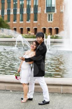 Vertical photo of young adult couple relaxing and holding each other with water fountain, flowers, trees and brick building in background 