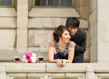 Horizontal photo of young adult couple, man behind his lady with his hands on her shoulders, looking over a balcony 