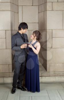 Vertical photo of young adult couple sharing a drink, as they look at each other, with stone wall in background 