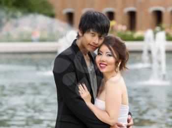 Closeup horizontal photo of young adult couple, looking forward, while holding each other with water fountain, flowers, trees and brick building in background 