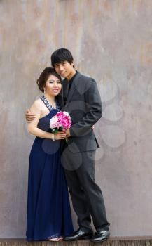 Vertical photo of young adult couple holding a bouquet of flowers, in between them, as they hug each other with rustic metal wall in background 

