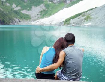 Photo of young adult lovers looking at pristine aqua color Mountain Lake with snow and woods in background 