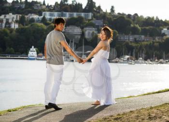 Horizontal photo of young adult couple, looking back, walking on pathway with harbor, reflecting sunlight and boats in Background