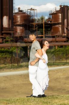 Vertical photo of young adult couple, locking arms while back to back, with factory and sky in background 