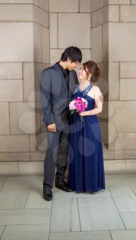 Vertical photo of young adult couple holding each other, with bouquet of flowers, as they look at each other, with stone wall in background 
