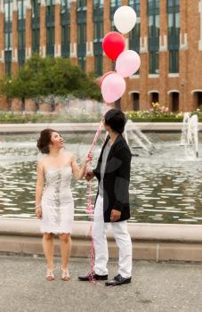 Vertical photo of young adult couple looking at several balloons overhead of them with water fountain, flowers, trees and brick building in background 