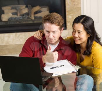 Photo of close mature couple looking at information, man giving thumbs up, on the computer screen together with fireplace in background  