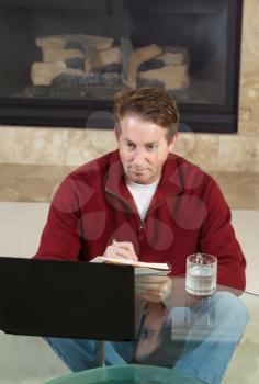 Vertical photo of mature man writing down notes from computer screen with fireplace in background 
