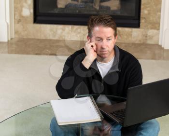 Photo of mature man, sitting down at glass table, working from home, looking at computer screen with fireplace and living room wall in background 
