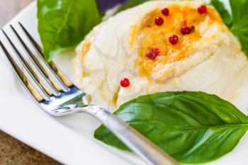 Closeup horizontal photo of baked stuffed sole fish, red peppercorn, sweet basil, fork inside white square plate on natural stone counter top 