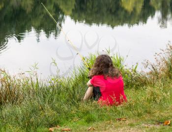Photo of young girl patiently waiting for fish to bite with lake and trees in background 