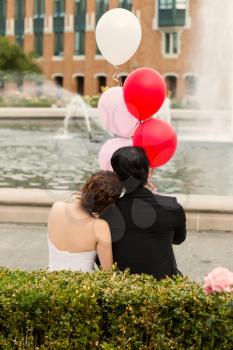 Vertical photo of young adult couple holding balloons while looking at water fountain with trees, flowers in background 