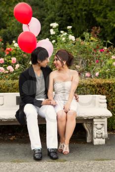 Vertical photo of young adult couple looking at each other while sitting down on a bench with several balloons, flowers and trees in the background 