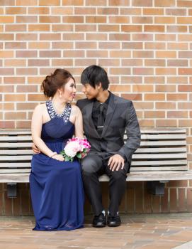 Vertical photo of young adult couple sitting on bench while looking at each other with brick wall in background 