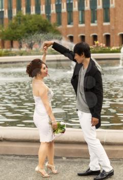 Vertical photo of young adult couple, doing a twirl, while holding hands with water fountain, flowers, trees and brick building in background 