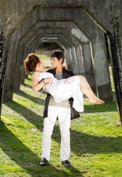 Vertical photo of young adult man lifting his lady in his arms while underneath a structured column wall in background 