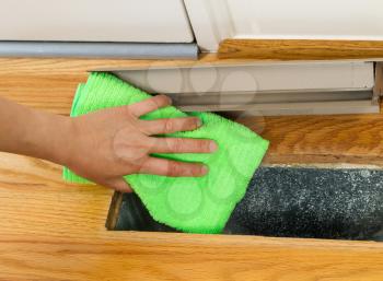 Horizontal photo of female hand cleaning, using microfiber rag, underneath grill plate of heater floor vent with Red Oak Floors and front door in background 