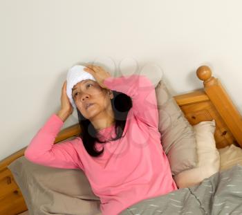 Photo of mature woman treating fever by holding wash cloth to her forehead while lying in bed 
