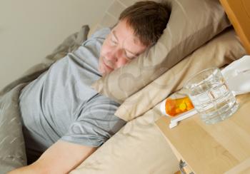 Closeup photo of medicine, thermometer, glass of water and tissues on night stand with mature man bedridden in background 