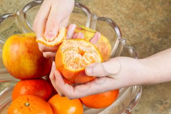 Horizontal photo of female hands peeling fresh ripe orange fruit from glass bowl filled with oranges and apples