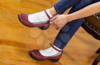 Horizontal photo of woman putting on causal shoes while sitting on leather padded footstool with red oak floors in background 