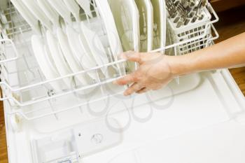 Horizontal photo of female hand closing lower dishwasher rack that is filled with white plates, stainless steel spoons and forks with red oak floors in background  