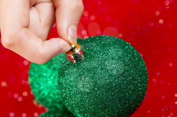 Horizontal photo of female fingers picking up green Christmas ornament against red background 