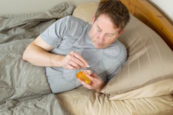 Horizontal photo of mature man looking at pill out of medicine container while lying in bed 