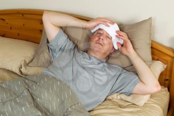 Horizontal photo of mature man treating fever by holding wash cloth to his forehead while lying in bed 